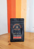 A bag of Mescalito Coffee’s Night Rider Blend Whole Coffee Beans rests on a wooden table before a wall with bold orange and yellow stripes.